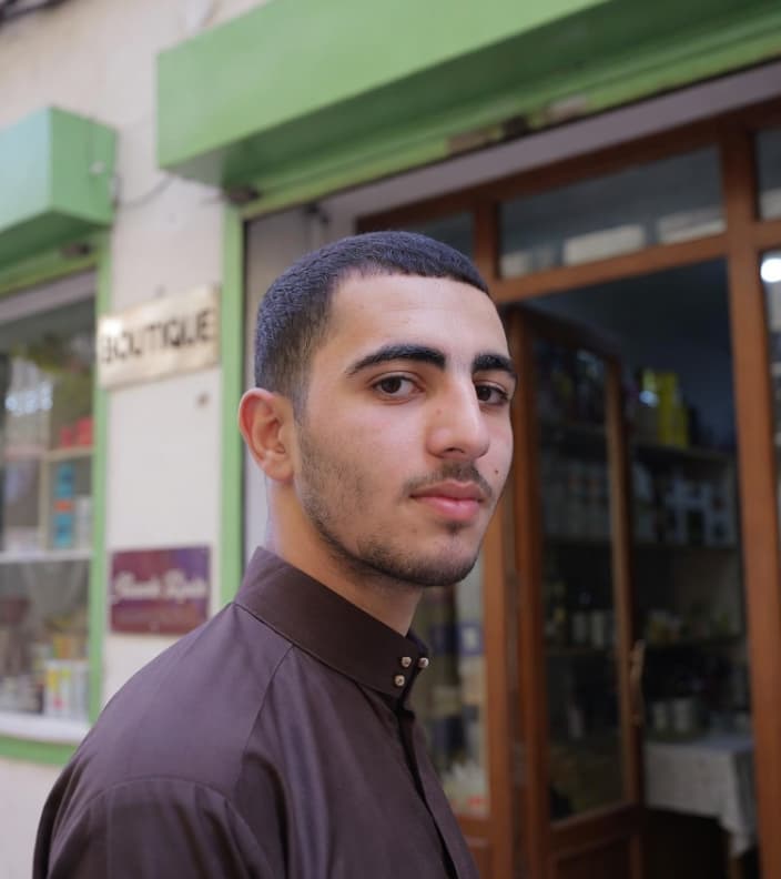 شاب بشعر قصير ولحية خفيفة يرتدي قميصاً بنياً ويقف أمام متجر في الشارع.
