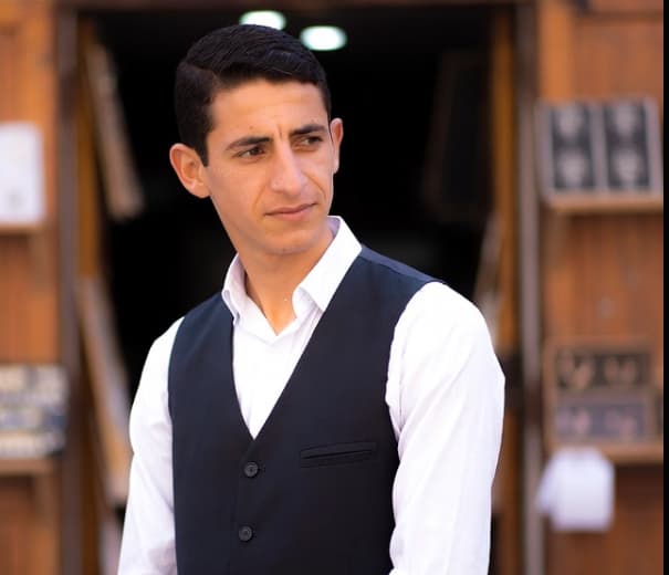 شاب يرتدي قميصاً أبيض وصديرية سوداء ينظر إلى الجانب أمام خلفية خشبية دافئة غير واضحة.