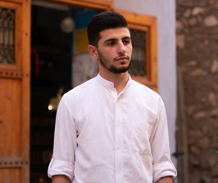 شاب ذو شعر داكن ولحية يرتدي قميصاً أبيض ويقف أمام باب خشبي كبير.