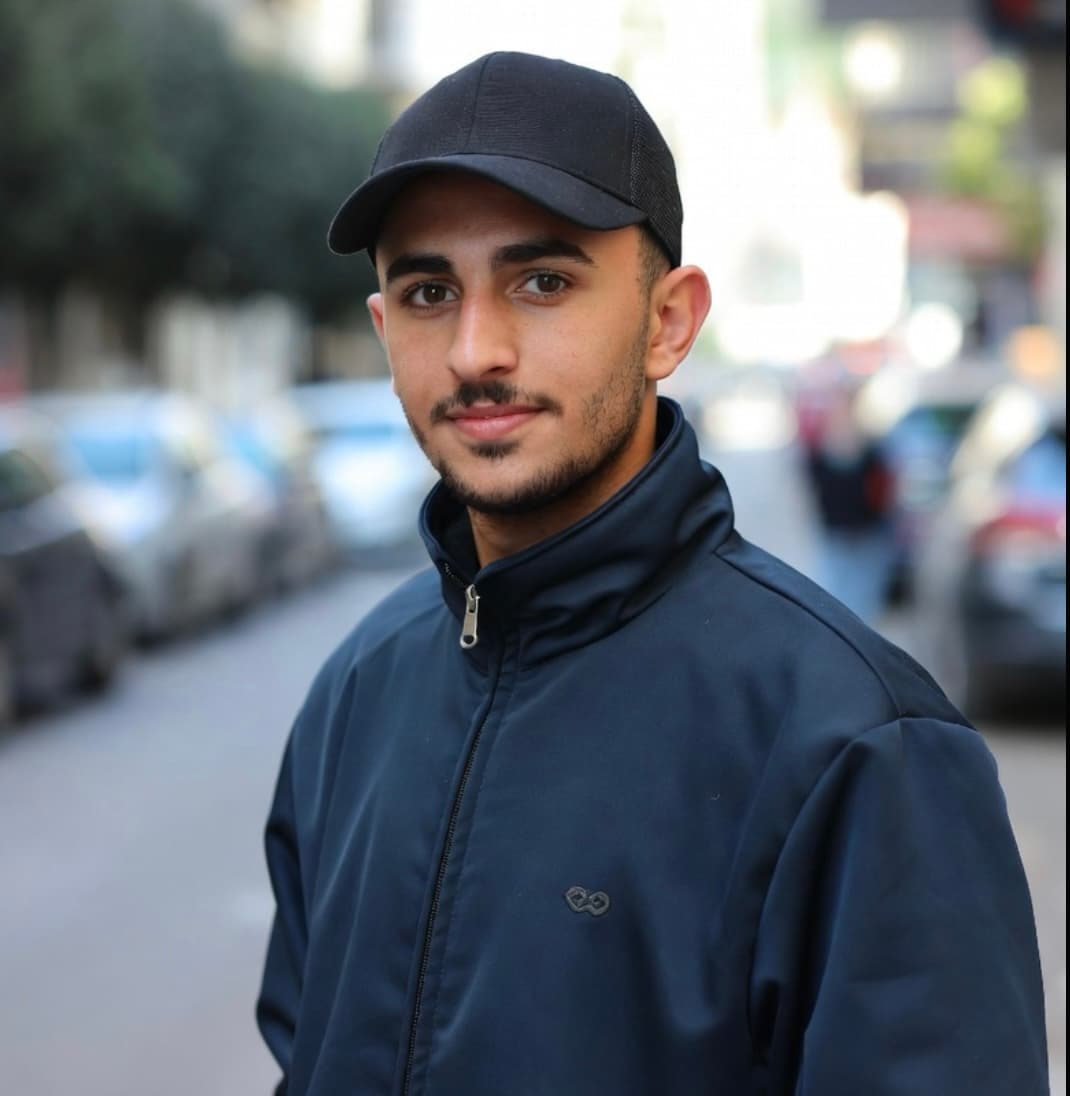 شاب ذو لحية خفيفة يرتدي قبعة سوداء وسترة زرقاء داكنة في شارع بالمدينة.