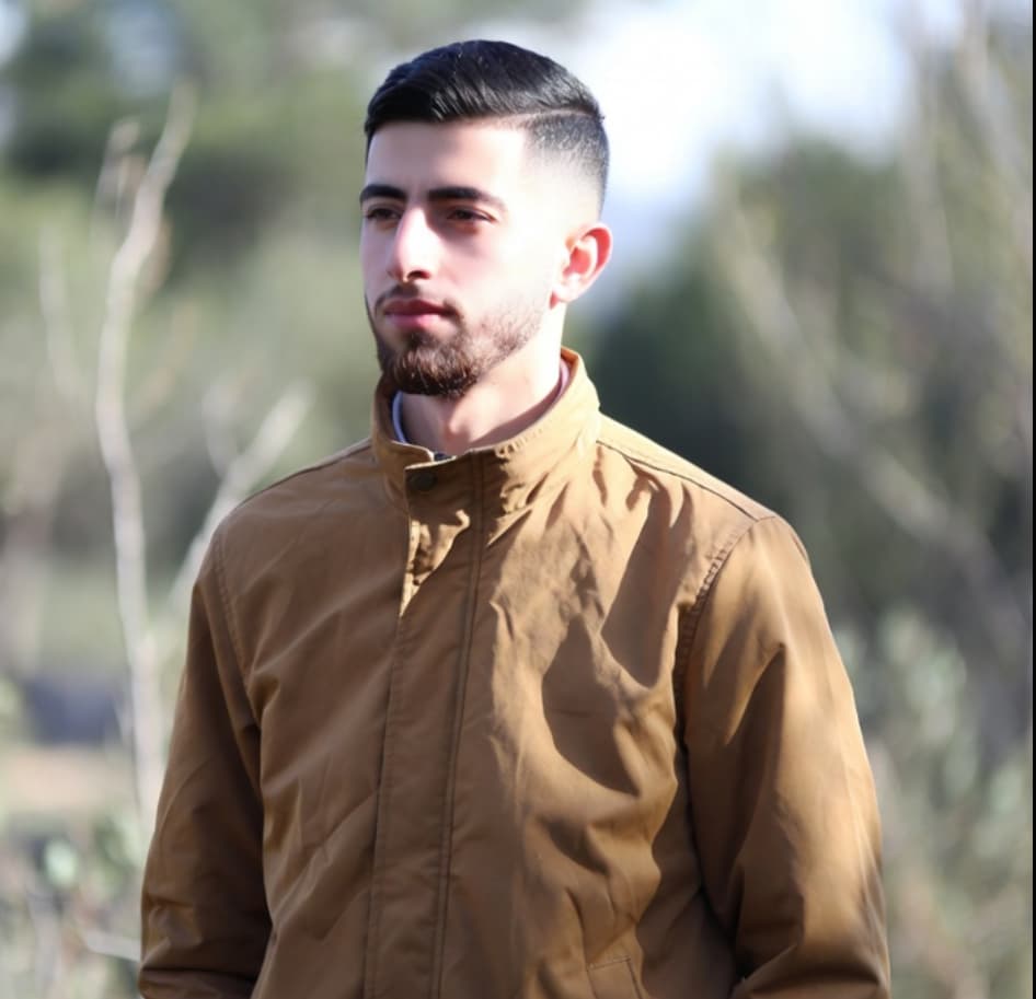 شاب بشعر داكن قصير ولحية يرتدي سترة بنية اللون في الهواء الطلق.